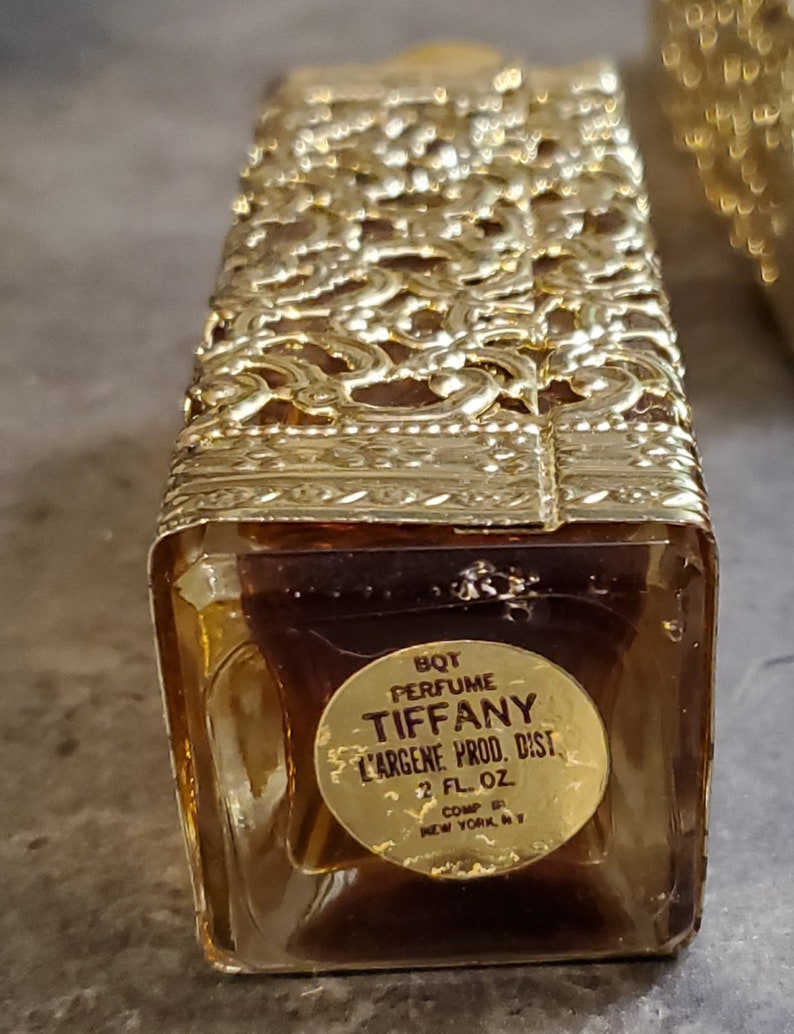 May include: A gold-colored glass bottle of Tiffany perfume with a gold label that reads "BQT PERFUME TIFFANY L'ARGENE PROD. DIST 2 FL. OZ COMP NEW YORK NY".