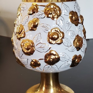 May include: A white ceramic vase with a gold rim and gold floral design. The vase has a round body and a wide base.
