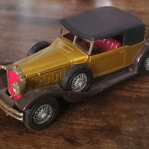 May include: A gold toy car with a black convertible top. The car has a red interior and chrome accents. The car is on a wooden surface.