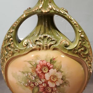 May include: A green and gold ceramic vase with a floral design. The vase has two handles and a round body. The floral design features pink and white flowers with green leaves.