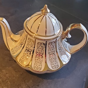 May include: A white teapot with a gold lid and intricate gold designs. The teapot has a rounded body and a curved spout.