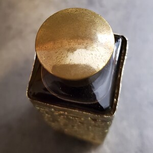 May include: A square glass bottle with a gold-colored lid and a textured gold-colored base. The bottle is filled with a dark liquid.