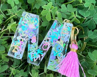 Puede incluir: Un llavero de resina transparente con la letra "M" incrustada con confeti de colores y un borla rosa.