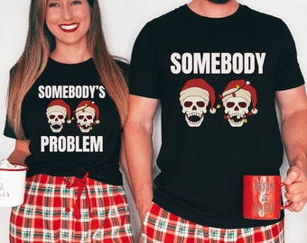 May include: A couple wearing matching black t-shirts with a skull and Christmas lights design. The woman's shirt says "Somebody's Problem" and the man's shirt says "Somebody". They are both wearing red and green plaid pyjamas.