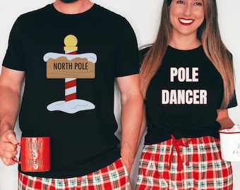 May include: A couple wearing matching black t-shirts with a Christmas theme. The man's shirt has a graphic of a North Pole sign with a candy cane pole. The woman's shirt says "POLE DANCER". They are both wearing red and green plaid pajama pants.