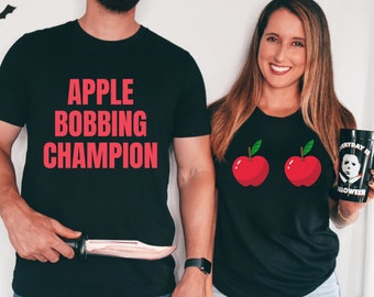 May include: A couple wearing matching black t-shirts with a Halloween theme. The man's shirt says "Apple Bobbing Champion" in red text. The woman's shirt has two red apples with green leaves on the front.