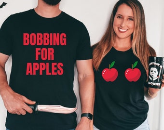 May include: A couple wearing matching black t-shirts with a Halloween theme. The man's shirt says "Bobbing for Apples" in red text. The woman's shirt has two red apples with green leaves on it.