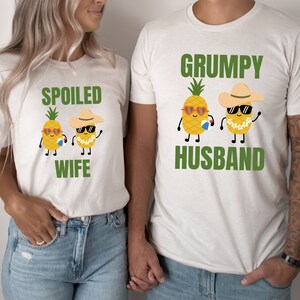 May include: Two white t-shirts with pineapple graphics and text. The first shirt says "Spoiled Wife" with a pineapple wearing sunglasses and a lei. The second shirt says "Grumpy Husband" with a pineapple wearing sunglasses and a hat.