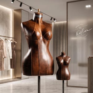 May include: Two brown leather-textured dress forms on metal stands. The larger form features a silver plaque on the neck and a base plate labeled "KARLOVADESIGN 2024". The background includes a clothing rack with garments and a glass wall with "ORIGINAL SINCE 2024 Karlova PROTOTYPE" text.