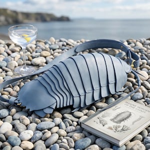 May include: A light blue shoulder bag shaped like a segmented marine isopod with a long adjustable strap, gold-toned metal hardware, and a smooth matte finish, accompanied by a book titled Marine Isopods of the Coast and a glass of water.