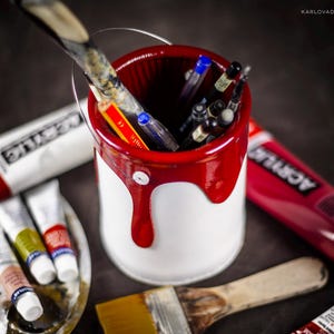 Peut inclure: Un pot de peinture blanc avec un bord rouge et de la peinture rouge qui coule sur les c&ocirc;t&eacute;s. Le pot est rempli de fournitures artistiques, notamment des pinceaux, des crayons et des stylos. Le pot est pos&eacute; sur une surface sombre avec d'autres fournitures artistiques.