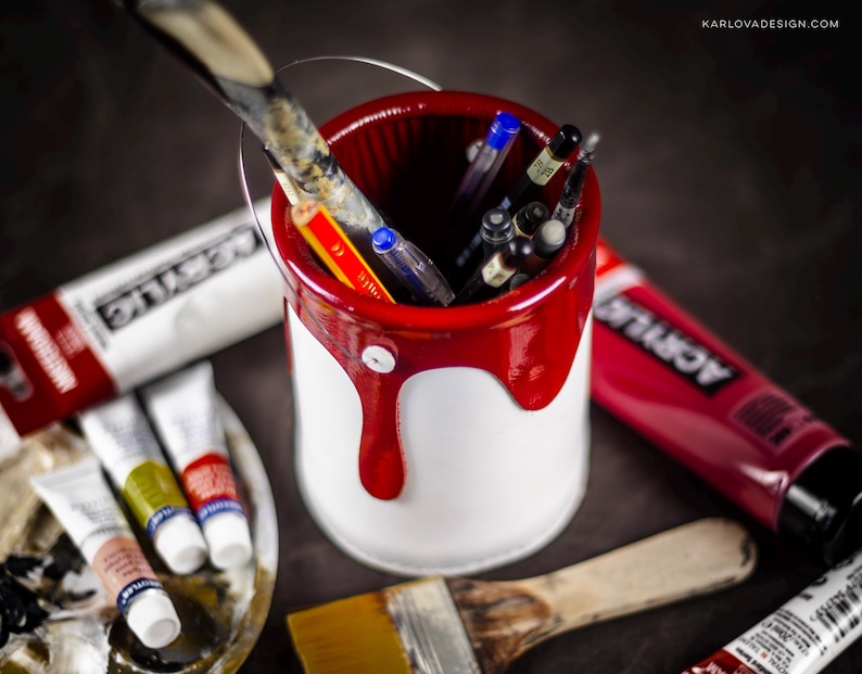 Peut inclure: Un pot de peinture blanc avec un bord rouge et de la peinture rouge qui coule sur les c&ocirc;t&eacute;s. Le pot est rempli de fournitures artistiques, notamment des pinceaux, des crayons et des stylos. Le pot est pos&eacute; sur une surface sombre avec d'autres fournitures artistiques.
