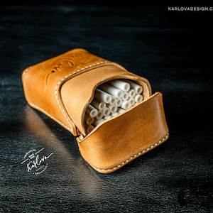 May include: A tan leather cigarette case with visible white stitching and a flap closure. The case is filled with white cylindrical items. The leather features an embossed logo and the text KARLOVADESIGN.COM is visible in the corner.
