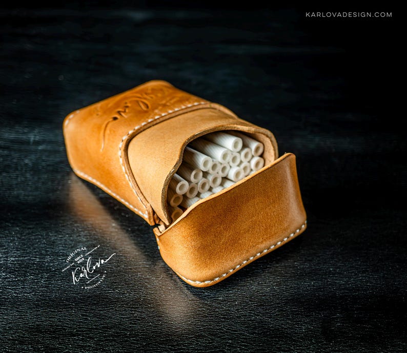 May include: A tan leather cigarette case with visible white stitching and a flap closure. The case is filled with white cylindrical items. The leather features an embossed logo and the text KARLOVADESIGN.COM is visible in the corner.