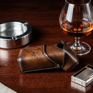 May include: A dark brown leather case with a visible embossed signature, positioned next to a silver metal ashtray, a glass of amber liquid, and a rectangular silver metal lighter with a textured grid pattern on its front surface.