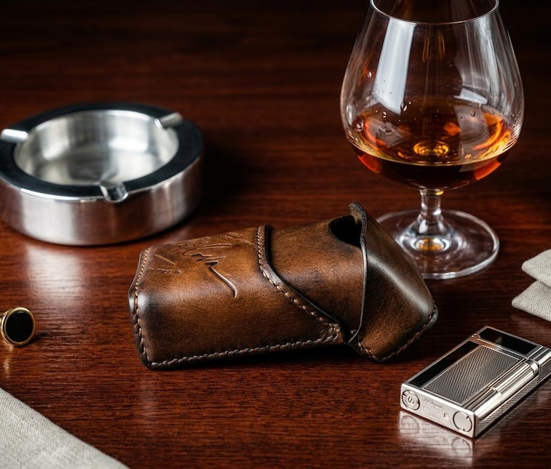 May include: A dark brown leather case with a visible embossed signature, positioned next to a silver metal ashtray, a glass of amber liquid, and a rectangular silver metal lighter with a textured grid pattern on its front surface.