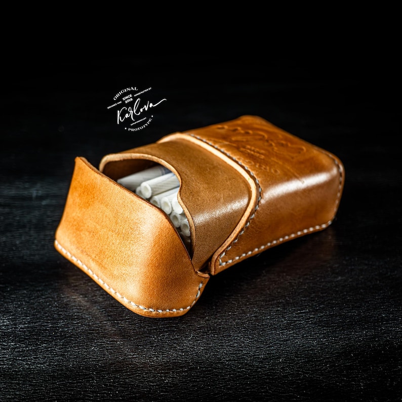May include: A tan leather cigarette case with white stitching, featuring a flap closure and visible white cigarette filters inside. The leather is embossed with the text Karlova Original Since 2016 Prototype.