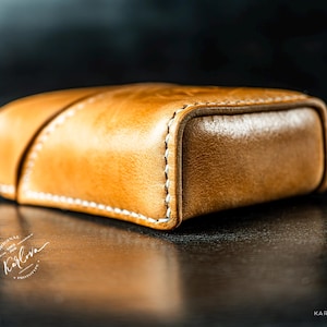 May include: A tan leather case with a smooth, polished finish and visible white stitching along the edges. The rectangular object features a diagonal seam across the top surface and includes the text Karlova and Karlovadesign.com.