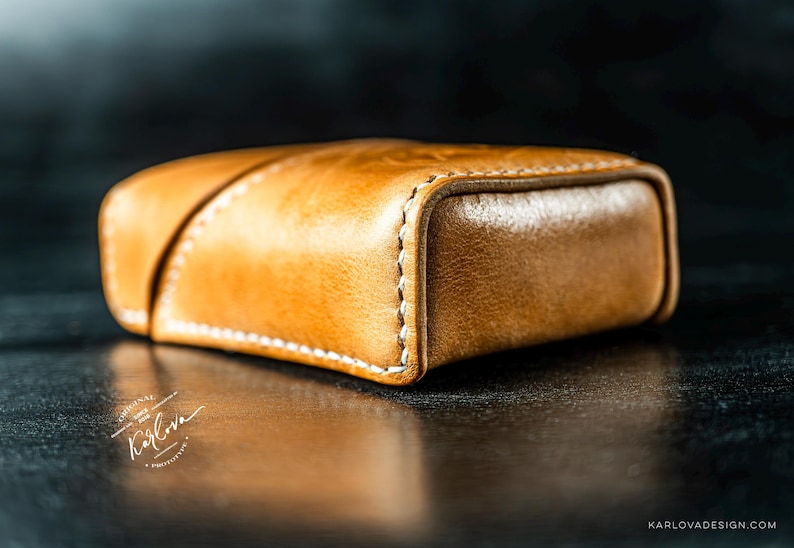 May include: A tan leather case with a smooth, polished finish and visible white stitching along the edges. The rectangular object features a diagonal seam across the top surface and includes the text Karlova and Karlovadesign.com.