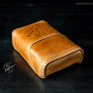 May include: A tan leather case with white stitching and a curved flap closure. The leather features a debossed cursive inscription on the top surface. The text KARLOVADESIGN.COM is visible in the upper right corner, and a circular logo with the text Karlova is in the lower left.