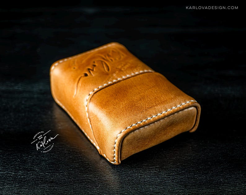 May include: A tan leather case with white stitching and a curved flap closure. The leather features a debossed cursive inscription on the top surface. The text KARLOVADESIGN.COM is visible in the upper right corner, and a circular logo with the text Karlova is in the lower left.