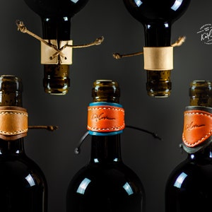 Puede incluir: Cinco botellas de vino con tapones de botella de cuero. Los tapones son de diferentes colores y tienen una firma manuscrita. Las botellas son todas de vidrio oscuro y tienen un líquido oscuro en su interior.