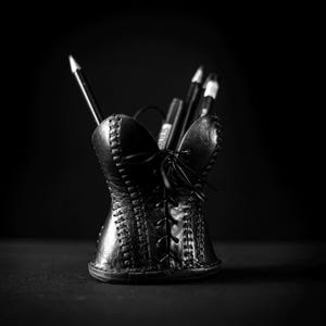 May include: A black pencil holder shaped like a corset with a lace-up front design and visible stitching details. The holder contains several dark pencils and pens, standing upright against a dark background, creating a monochromatic aesthetic.