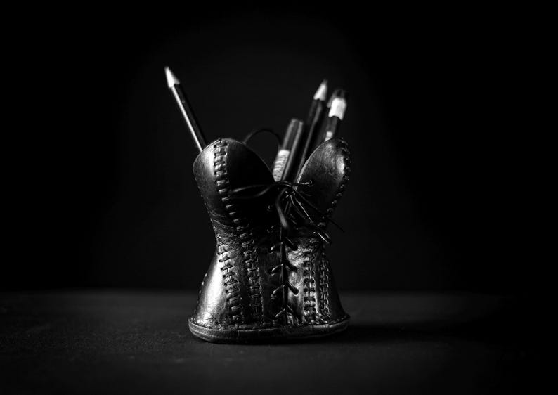 May include: A black pencil holder shaped like a corset with a lace-up front design and visible stitching details. The holder contains several dark pencils and pens, standing upright against a dark background, creating a monochromatic aesthetic.