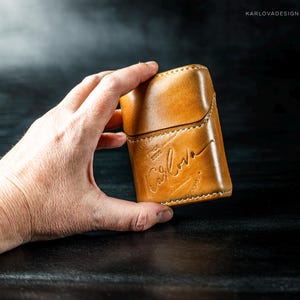 May include: A tan leather case with a flap closure and visible white stitching. The front is embossed with the text "KARLOVA" in a cursive script, along with the smaller text "SINCE 2020" positioned above the brand name on the smooth surface.