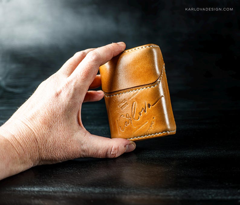 May include: A tan leather case with a flap closure and visible white stitching. The front is embossed with the text "KARLOVA" in a cursive script, along with the smaller text "SINCE 2020" positioned above the brand name on the smooth surface.