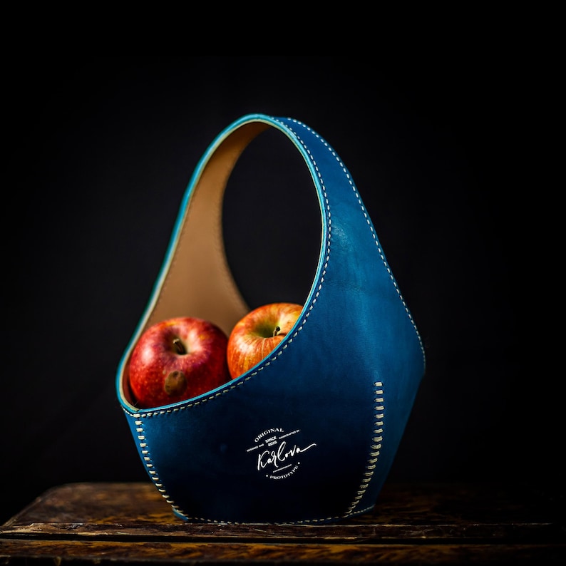 May include: A deep blue leather basket with a single arched handle and light-colored stitching along the edges. The basket contains two red apples. The front features a white logo with the text Original Since 2018 Karlova Prototype.