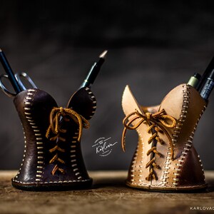 May include: Two leather pencil holders shaped like corsets, one dark brown and one light tan. Each features white contrast stitching, a front lace-up design with brown leather cords, and holds various pens and pencils. Text reads Karlova.