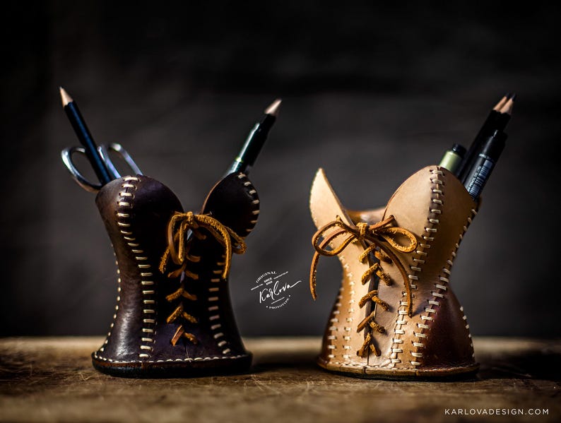 May include: Two leather pencil holders shaped like corsets, one dark brown and one light tan. Each features white contrast stitching, a front lace-up design with brown leather cords, and holds various pens and pencils. Text reads Karlova.