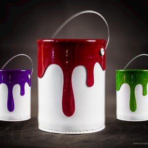 Peut inclure: Trois contenants en forme de pot de peinture avec des poign&eacute;es. Les contenants sont blancs avec de la peinture rouge, violette et verte qui coule sur les c&ocirc;t&eacute;s.