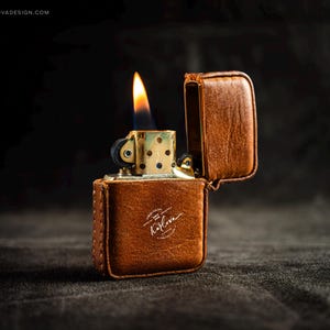May include: A brown leather lighter case with a gold-colored lighter inside, with a flame lit. The case has a stitched design and a logo that reads "Karlova Design". The lighter is open, and the flame is bright.
