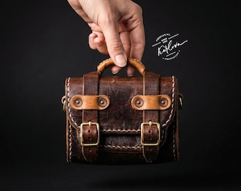 May include: A dark brown leather satchel with a top handle, featuring two front straps with brass buckles and rivets. The bag has visible white stitching along the edges and a logo printed in white text that reads Original Karlova Prototype.