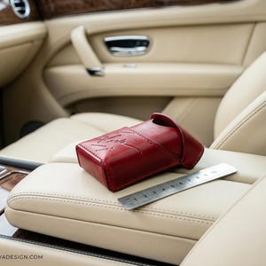 May include: A deep red leather case with a flap closure and a debossed signature logo on the front. A silver metal ruler is placed next to the case, showing a length of approximately 10 cm for the item. The case has visible stitching.