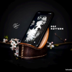 Puede incluir: Un soporte de teléfono de cuero marrón con un diseño cosido. El soporte sostiene un teléfono inteligente negro con un perro blanco en la pantalla. La hora en el teléfono es 17:48. El texto "PDF PATTERN" es visible en la imagen.