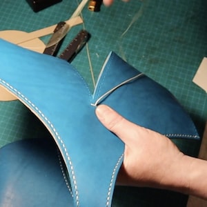 May include: A bright blue leather piece being hand-stitched with thick white thread. The leather has a smooth matte finish and is folded into a V-shape. The stitching follows the curved edges of the blue material, creating a visible seam.