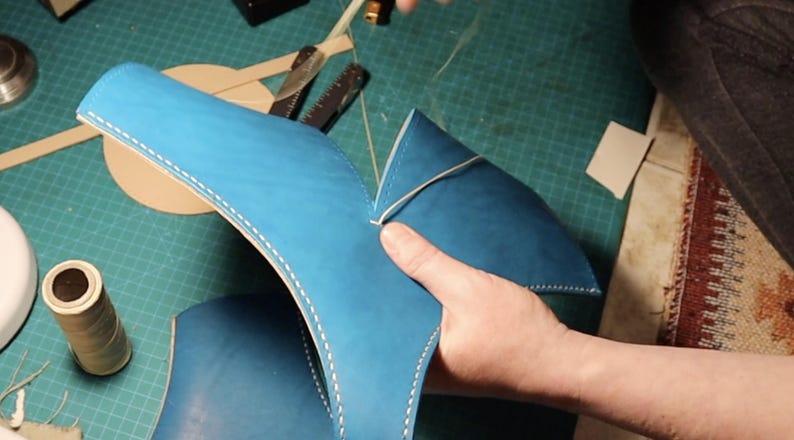 May include: A bright blue leather piece being hand-stitched with thick white thread. The leather has a smooth matte finish and is folded into a V-shape. The stitching follows the curved edges of the blue material, creating a visible seam.