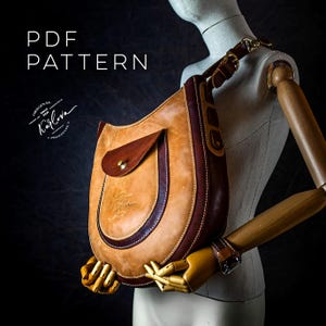以下が含まれることがあります： フラップとポケットが付いた茶色の革製のバッグのPDFパターン。バッグにはストラップが付いており、木製のハンドで持たれています。