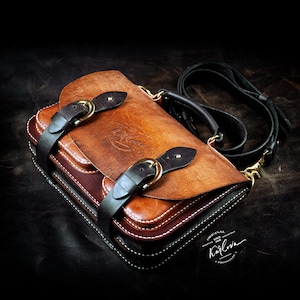 Puede incluir: Un maletín de cuero estructurado con una solapa color canela, dos correas con hebilla marrón oscuro, un asa negra y una correa de hombro extraíble. El bolso presenta costuras blancas en contraste y un logotipo estampado Karlova Original.