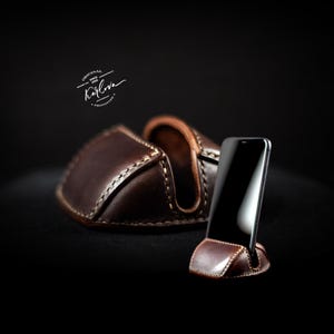 Può includere: Un supporto per telefono in pelle marrone scuro con cuciture chiare visibili, caratterizzato da un design curvo per mantenere lo smartphone in posizione verticale. La pelle ha una finitura liscia e un logo con il testo Original Karlova Prototype.