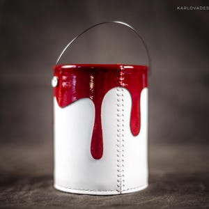 Peut inclure: Un pot de peinture blanc avec un couvercle rouge et de la peinture rouge qui coule sur les c&ocirc;t&eacute;s. Le pot a une poign&eacute;e en m&eacute;tal et est en cuir.