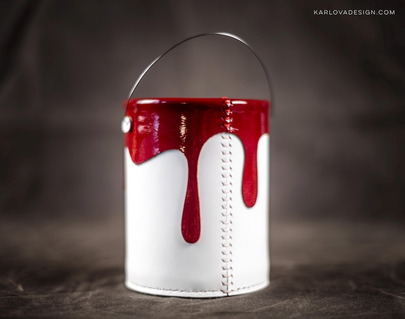 Peut inclure: Un pot de peinture blanc avec un couvercle rouge et de la peinture rouge qui coule sur les c&ocirc;t&eacute;s. Le pot a une poign&eacute;e en m&eacute;tal et est en cuir.