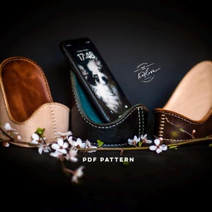 Puede incluir: Dos soportes de teléfono de cuero marrón con un soporte de teléfono de cuero negro en el centro. El soporte de teléfono del centro tiene un teléfono inteligente negro dentro. La pantalla del teléfono muestra la hora 17:48. Los soportes de teléfono están sobre un fondo oscuro con flores blancas.