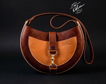 Può includere: Una borsa a tracolla in pelle a forma di mezzaluna nei toni del marrone scuro e cuoio, con cinturino centrale verticale, chiusura a moschettone in ottone e tracolla lunga. Presenta una tasca frontale color cuoio con logo e cuciture bianche.