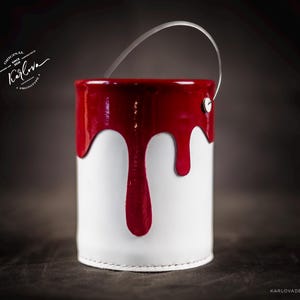 Peut inclure: Un pot de peinture blanc avec un couvercle rouge et de la peinture rouge qui coule sur le c&ocirc;t&eacute;. Le pot a une poign&eacute;e en m&eacute;tal.