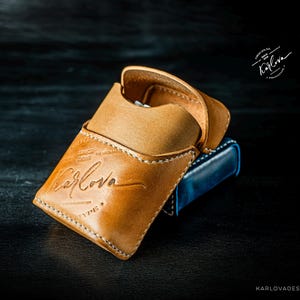 May include: A tan leather case with a flap closure and white stitching. The front is embossed with the text Karlova, Since 2016, and Type. A blue object is partially visible underneath the tan case. The logo Karlova is also printed in the top right.