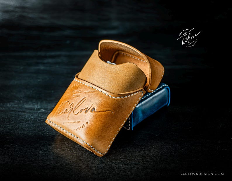 May include: A tan leather case with a flap closure and white stitching. The front is embossed with the text Karlova, Since 2016, and Type. A blue object is partially visible underneath the tan case. The logo Karlova is also printed in the top right.